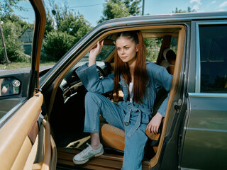 Wall Mural - Stylish young woman in denim outfit sitting in a car, exuding confidence and joy in a sunny outdoor setting with greenery.