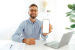 © Kateryna - Positive successful businessman with glasses sitting at a desk in the office and showing a smartphone with a blank mock-up screen. Mockup display concept for apps or advertising, presentation