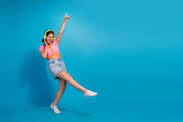 Wall Mural - Young woman dancing joyfully with headphones, wearing trendy summer outfit against blue background