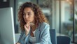© natakot - A focused woman with curly hair sits at a modern desk, deep in thought as she analyzes data on her computer screen. Her professional attire and contemplative expression convey determination and