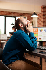 Wall Mural - Young male employee seated at office desk, rubbing his face from exhaustion. Businessman looking up, feeling tired and overwhelmed as desktop monitor displays startup company analysis.