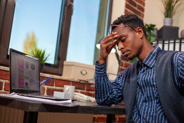 Wall Mural - African American entrepreneur sits tiredly at desk, holding his head as he analyzes business project plans. Black male manager looking drained after demanding day at work in startup office.