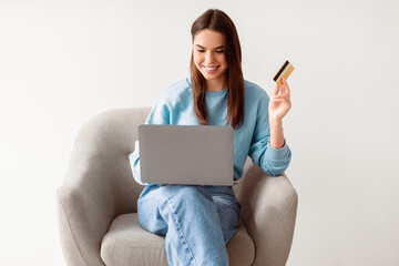 Wall Mural - Attractive young woman with credit card and laptop sitting in cozy armchair, ordering goods on internet, buying things in web store, making contactless payment. Online shopping, remote banking