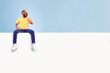 © Prostock-studio - Happy African American man sitting on white blank placard, shouting, announcing huge sale on blue studio background, mockup for logo or design. Positive guy with empty advertising banner, free space