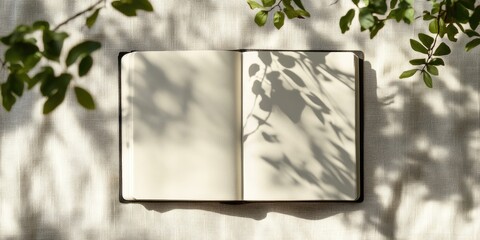 Canvas Print - Blank open notebook on a light background surrounded by green leaves casting soft shadows, creating a serene and tranquil atmosphere.