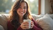 © natakot - A cheerful young woman with long, wavy hair sits comfortably on a cozy couch, holding a steaming cup of coffee. Her radiant smile and relaxed posture create a warm, inviting atmosphere, perfect for a