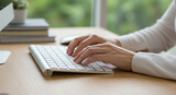 Manos tecleando en un teclado blanco sobre un escritorio de madera. Fondo con libros y una planta, enfoque en la acción de escribir.