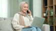 © lina - smiling woman in hijab working from home using laptop and phone