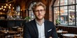 © Imagine Crane - A well-dressed young man stands with arms crossed in a lively restaurant filled with tables and warm lighting. Large windows allow natural light to illuminate the space beautifully