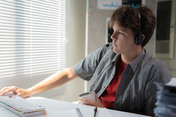 Young student in casual attire focuses on schoolwork while wearing headphones at a bright desk setting. Natural light streams through nearby window blinds creating a peaceful study environment