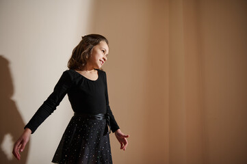 Wall Mural - Young girl in a black dress posing gracefully in a dimly lit room surrounded by soft shadows.