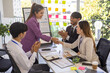 © Pattra - A group of people are gathered around a white board with a woman giving a presentation. Scene is positive and collaborative, as the group is clapping and smiling