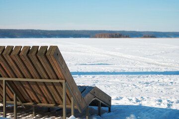 Wall Mural - Winter landscape. Fragment of beach wooden recliner on the background of frozen river covered with snow. Copy space.