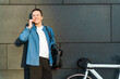 © muse studio - Young man smiles while talking on the phone beside his bicycle against a modern building wall