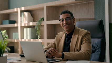 Happy African American man ethnic male entrepreneur business leader businessman employee ceo manager working on laptop computer typing online project at office looking at camera smiling cheerful smile