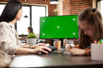 Caucasian mother using desktop pc with greenscreen as little girl sits nearby at desk, in brick wall office. Woman working on computer having isolated chromakey display as female child draws at table.