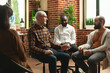 © DC Studio - Caucasian men having conversation as black man smiles at camera in brick wall room. Young and old male individuals sitting together and discussing about mental health at aa group meeting.