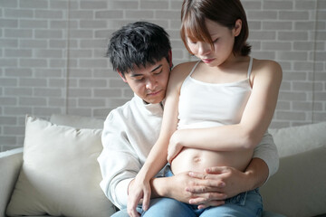 Wall Mural - A Japanese pregnant woman in her 20s, dressed to show her stomach, is sitting on a sofa in her apartment, her husband holding her, and the two of them looking at her stomach.