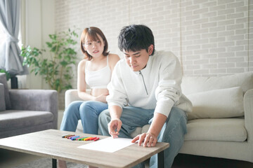 Wall Mural - A pregnant Japanese woman in her 20s, wearing clothes that reveal her stomach, and her husband are sitting on a sofa in their apartment, drawing a picture.