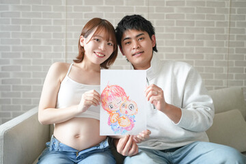 Wall Mural - A Japanese pregnant woman in her 20s, wearing clothes that show her stomach, and her husband sit on the sofa in their apartment and draw a picture.