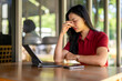 © Wasana - A woman in a red shirt is sitting at a table with a laptop and a cell phone
