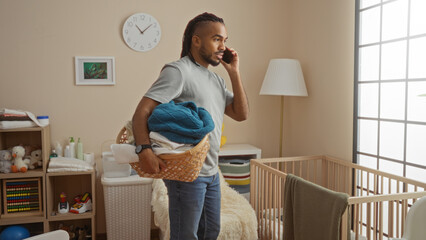 Young man with braids talking on phone while holding laundry basket in cozy bedroom with cradle and stylish decor, exuding comfort and modern family lifestyle.