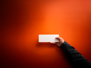 Aufkleber - A hand in dark sleeve holds a blank white rectangle against a vibrant orange backdrop.  The scene is simple yet striking with strong color contrast and soft lighting.