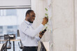© anatoliycherkas - Young african american manager taking notes on colorful sticky notes attached to a wall in a modern office, using a pen and consulting his smartphone