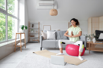 Wall Mural - Sporty African-American woman with laptop meditating on fitball at home