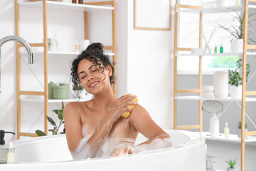 Wall Mural - Beautiful young happy African-American woman taking bath with sponge and soap foam at home