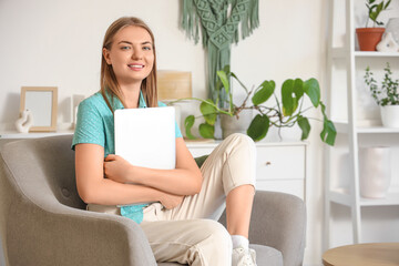 Wall Mural - Smiling young woman with laptop sitting in armchair at home