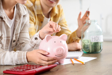 Wall Mural - Young couple putting coin into piggy bank at table in kitchen, closeup