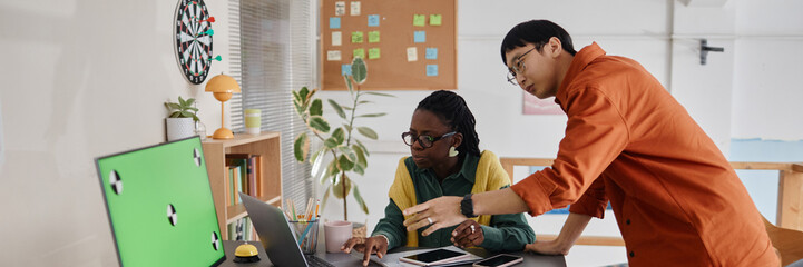 Two coworkers collaborating on project at desktop computer while discussing ideas surrounded by office decor and plants. Green screen visible on monitor in foreground