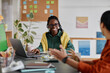 © Seventyfour - Smiling woman sitting in office environment with colleague, engaging in casual conversation. Books, laptop, and plant seen in background, providing a warm, professional setting