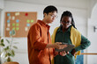 © Seventyfour - Asian man and African American woman standing in office while discussing together and holding tablet. Notice board, plant, and office interior visible in background