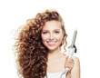 © artmim - Joyful woman with curly hairstyle using hot curling tongs iron, indoor fashion portrait. Coiffure, beauty salon and hair styling concept