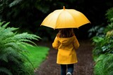 A little girl in a yellow raincoat holding a matching umbrella, walking on a rainy path surrounded by vibrant green foliage