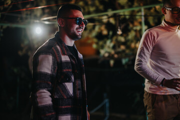 Sticker - A man wearing glasses and a plaid shirt smiles during a festive gathering outdoors under decorative lights.