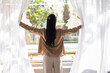 © fizkes - Rear view of young hotel guest woman drawing white veils at window in morning, waking up, looking at backyard, patio, enjoying fresh air, daylight, enjoying vacation, recreation