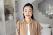 © fizkes - Serious beautiful young Vietnamese woman in casual posing for head shot, standing in white colored cozy home interior, looking at camera. Female Asian model front video call screen portrait