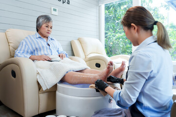 Wall Mural - Asian elderly woman receiving foot spa treatment from young spa therapist in uniform. Clean, serene spa setting emphasizes professional care, comfort, and relaxation.