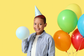 Wall Mural - Cute little African-American boy in party hat with air balloons celebrating birthday on yellow background