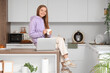 © Pixel-Shot - Young woman with cup of coffee and modern laptop sitting on table in kitchen