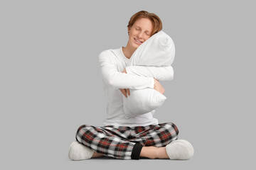 Wall Mural - Handsome young happy man in pajamas with pillow sitting on grey background