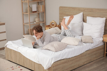 Wall Mural - Handsome young happy man in pajamas with pillows and laptop lying on bed at home