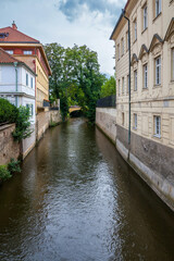 Wall Mural - A tranquil canal lined with historic buildings and lush trees reflects the cloudy sky, evoking a serene urban scene.