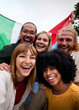 © CarlosBarquero - Vertical. Multi-ethnic group of young cheerful people taking a selfie while holding an italian flag and laughing, celebrating and having fun together in the city center