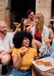 © CarlosBarquero - Vertical. Group of cheerful young multiethnic friends are laughing and enjoying eating street food pizza together while sitting on steps in front of a historical building