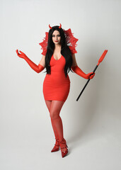 Wall Mural - portrait of beautiful dark haired model wearing elegant red dress with gothic fantasy burlesque halloween themed devil costume, with horns. isolated pose on white studio background