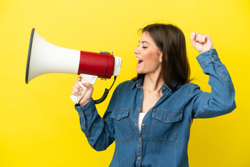 Wall Mural - Young caucasian woman isolated on yellow background shouting through a megaphone to announce something in lateral position